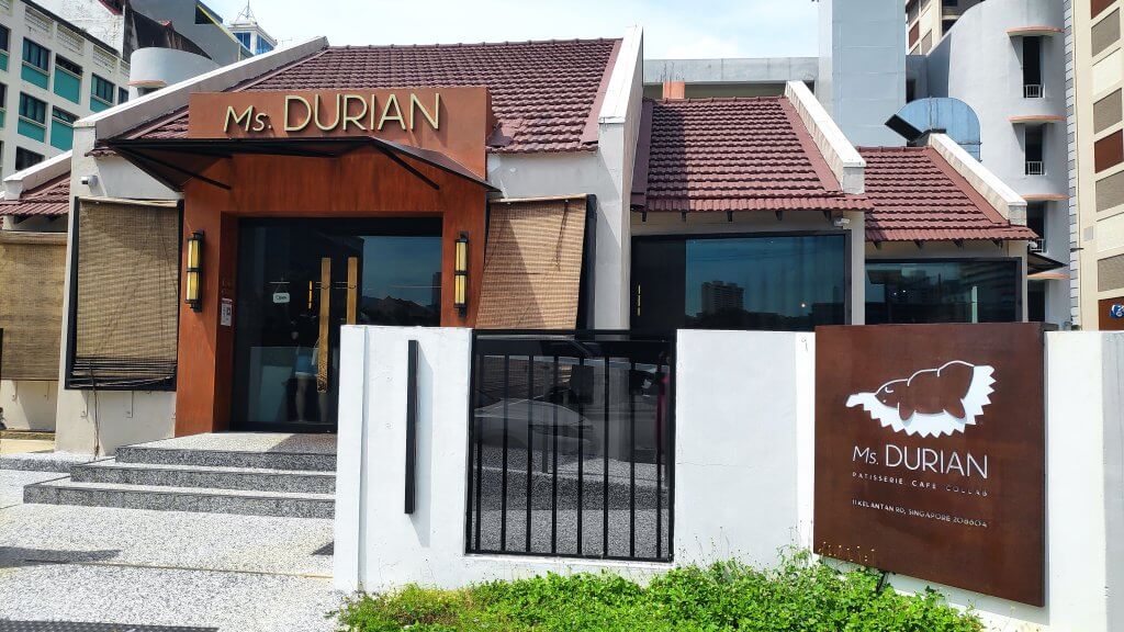 Image Description: Facade of Ms. Durian painted in brown with white fences and on the right side of the fence, brown signage with Ms. Durian and a durian logo. Above the entrance door with three steps stairs a signage says Ms Durian