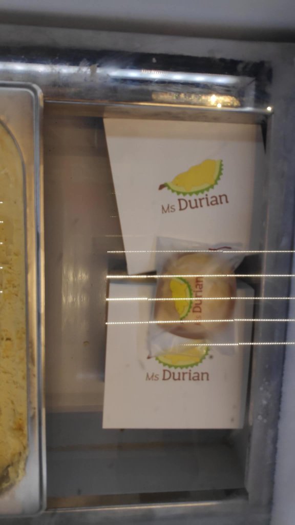 Image Description: Bun inside the plastic packaging and two paper packaging with durian logo in yellow and green. With text Ms Durian