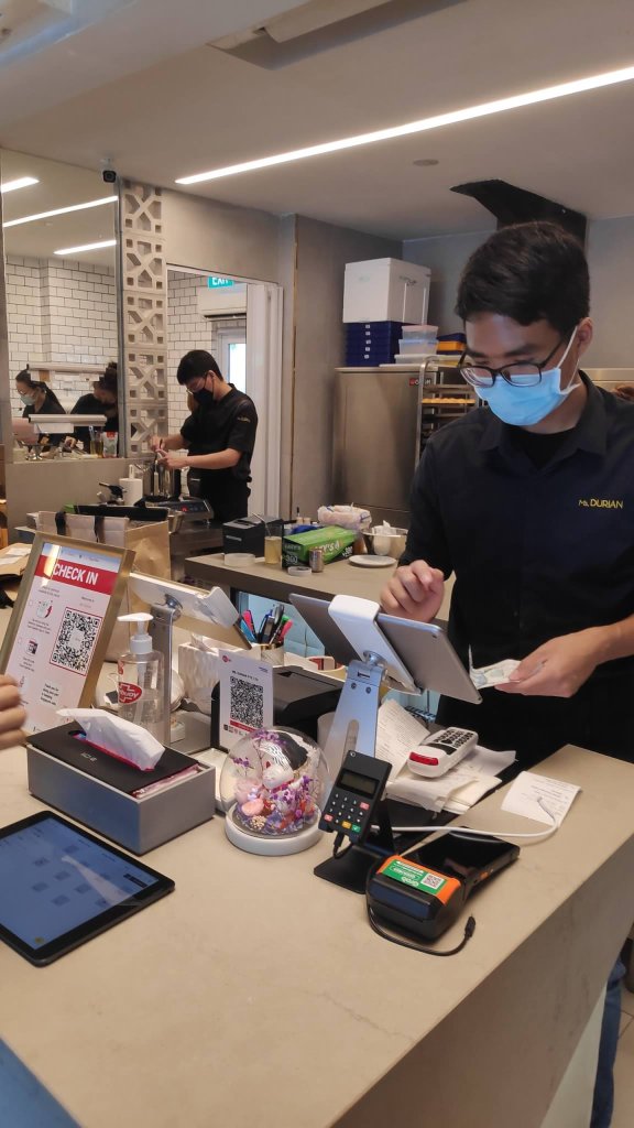 Image description: Men and women in black uniform with mask working inside the store counter. One accepting the orders and in front of cash register machine then the three staffs at the back are preparing
