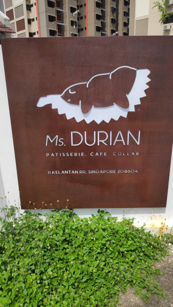 Image description: Signage painted in brown with durian. The durian logo is brown seeds and meats then the durian skin in white. Below the logo says Ms. Durian and small text with what they sell and address